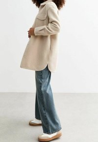 Camisa chaqueta de mezcla de lana color beige con un bolsillo, dobladillo redondeado, cierre de botones, combinada con pantalones vaqueros de pierna ancha color azul claro y zapatillas blancas.