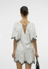 Top bianco a maniche corte con chiusura a cravatta sul retro e rifiniture in pizzo nero scallopato. Tessuto leggero; abbinato a pantaloni corti coordinati.