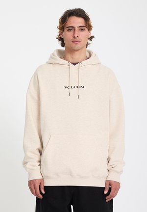 Jeune homme aux cheveux châtain clair portant un sweat à capuche beige Volcom avec une poche avant et un pantalon noir, debout devant un fond blanc uni.