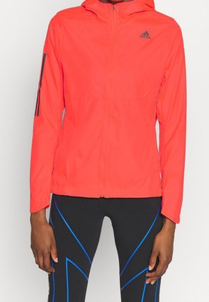 Personne portant une veste à capuche Adidas orange vif et des leggings noirs avec des lignes géométriques bleues.