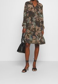 Sukienka z długim rękawem, z kołnierzykiem w ciemnozielonym kolorze z kwiatowym wzorem w brzoskwiniowym i niebieskim odcieniu, z falbaną na dole i luźnym krojem. Czarna torba na ramię.