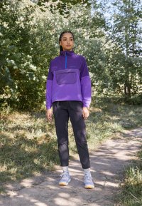 Lilla halv-zip jakke med en stor frontlomme, kombinert med svarte smale bukser og lyseblå sneakers i en naturlig setting.