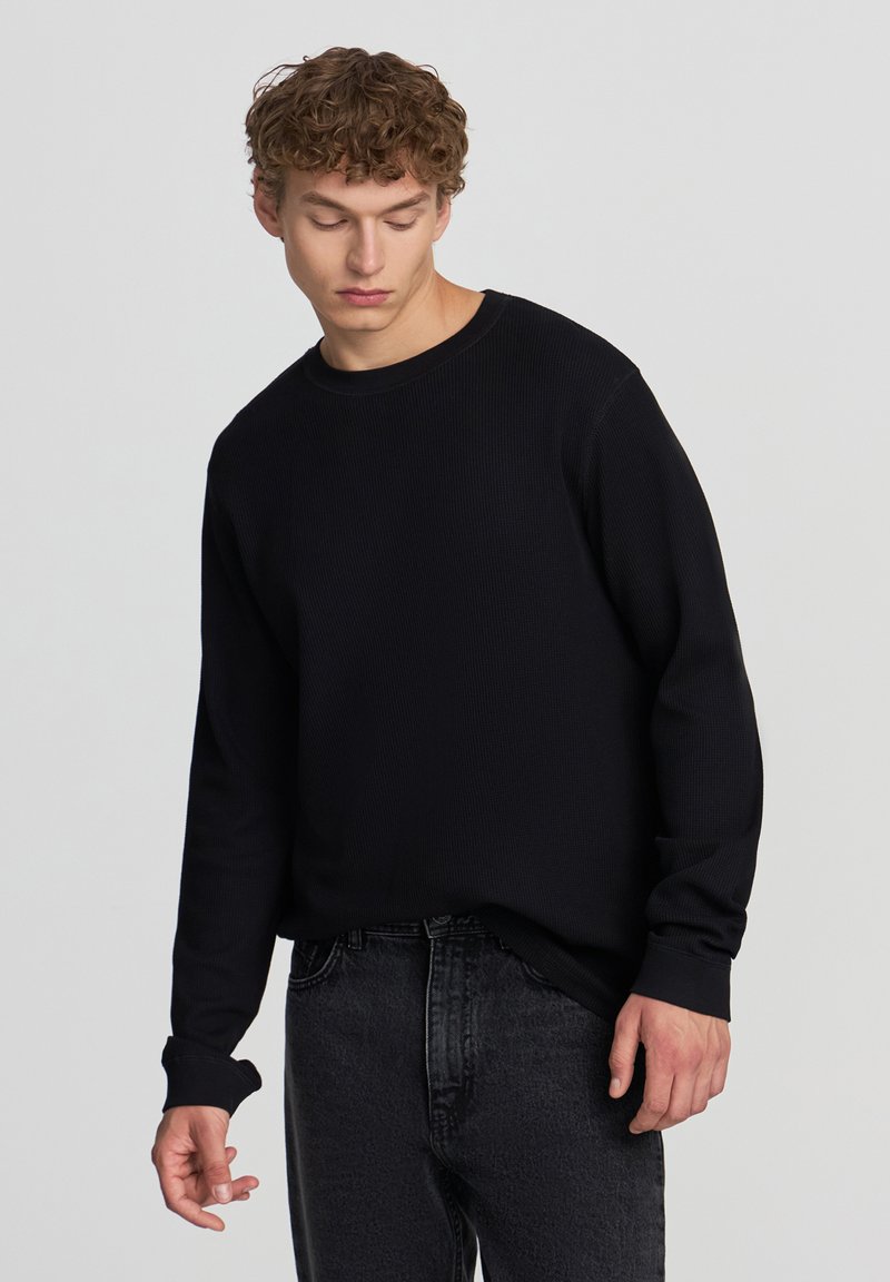 Giovane con capelli ricci che indossa una maglietta nera a maniche lunghe e jeans grigio scuro, che guarda in basso con un'espressione neutra.