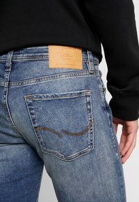 Blå denimjeans med en läderlogga, kontrasterande sömmar och rundade ficköppningar, sedd bakifrån på en svart topp.