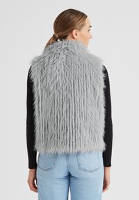 Graue Kunstpelz-Weste mit langem, flauschigem Material. Sie hat einen hohen Kragen, einen verkürzten Schnitt und wird mit einem schwarzen Langarmoberteil und blauen Jeans kombiniert.