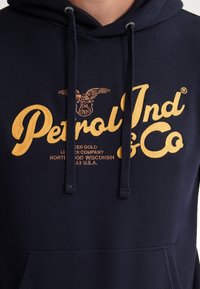 Marine hoodie met gele borduring. Beschikt over een logo met een arend, merknnaam "Petrol Ind & Co," en een voorzak. Verstelbare koordjes.