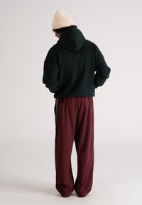 Felpa con cappuccio verde scuro dalla vestibilità ampia, abbinata a pantaloni della tuta bordeaux con tasche laterali e gamba leggermente affusolata. Berretto lavorato a maglia beige.