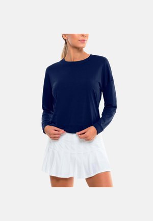 Frau trägt ein marineblaues Langarmoberteil und einen weißen plissierten Rock, steht mit den Händen am Saum des Oberteils.