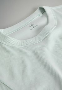 Gros plan sur un T-shirt col rond en tissu côtelé vert pâle avec une étiquette visible indiquant la taille M et fabriqué au Bangladesh.