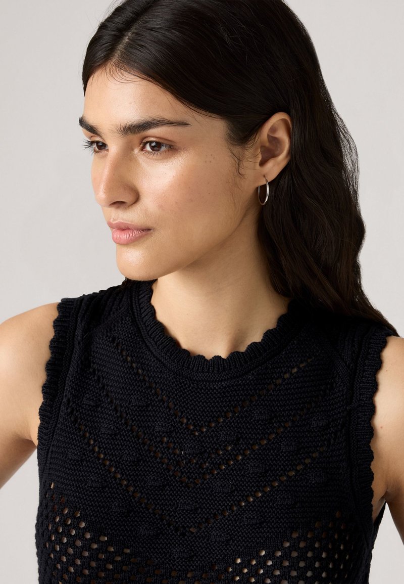 Kobieta z długimi ciemnymi włosami, nosząca bezramiowy czarny top dzianinowy z teksturowanymi wzorami i kolczykami w kształcie kół, spoglądająca w lewo.