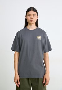 Młoda osoba z długimi ciemnymi włosami nosi szary t-shirt z małym kwiatowym logo na klatce piersiowej i oliwkowe spodnie, stoi na tle białej ściany.