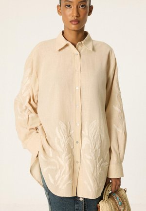 Vrouw draagt een losse beige blouse met knopen en geborduurde bladvormen, houdt een gevlochten mandtas vast, staat tegen een effen achtergrond.