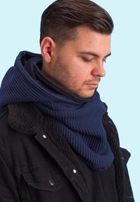 Navy blue knit scarf with textured pattern, layered for warmth, worn over a dark shearling coat. Smooth and thick fabric, casual fit.