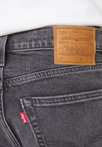 Dunkelgraue Jeans aus Denim mit strukturierter Oberfläche, ausgestattet mit einem Markenetikett aus cognacfarbenem Leder und einem roten Etikett an der Gesäßtasche.