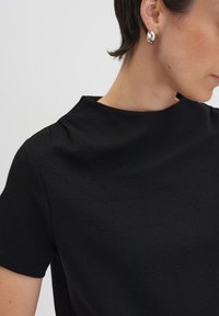 Zwarte korteblauwe top met een hoge hals, textuurstof en subtiele stikdetails, gecombineerd met een eenvoudige zilveren ringoorbel.