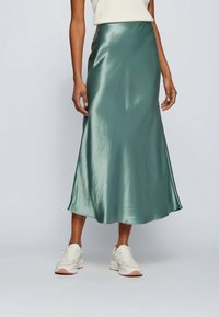 Satiini, keskipitkä hame pehmeässä vihreässä sävyssä, jossa on liehukkapuku, sileä rakenne ja hienovarainen kiilto, yhdistettynä valkoisiin lenkkikenkiin.