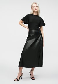 Femme blonde portant un haut noir à manches courtes avec des volants, une jupe en cuir midi noire et des sandales à talons hauts noires, debout.