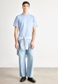 Világoskék rövid ujjú pamut póló, széles szárú világoskék farmernadrággal párosítva. Csíkos ing van megkötve a deréknál. Fekete cipő.