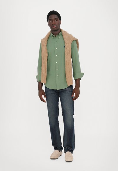 Chemise verte à boutons, pull en tricot beige drapé sur les épaules, jean foncé et mocassins clairs. Couleurs unies avec des détails subtils.