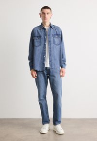 Denim shirt met twee borstzakken, blauwe kleur, knoopsluiting en lange mouwen, gecombineerd met blauwe jeans en witte schoenen.