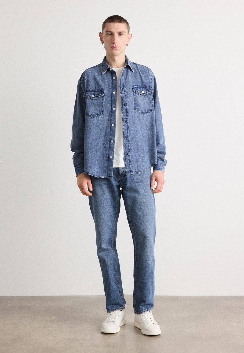 Jeanshemd mit zwei Brusttaschen, blauer Farbe, Knopfverschluss und langen Ärmeln, kombiniert mit blauen Jeans und weißen Schuhen.
