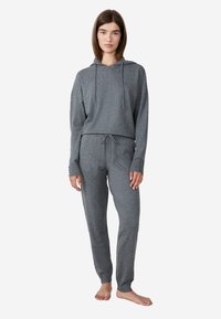 Vrouw die blootsvoets staat, gekleed in een grijze gebreide hoodie en bijpassende joggerbroek met trekkoord, tegen een effen achtergrond.