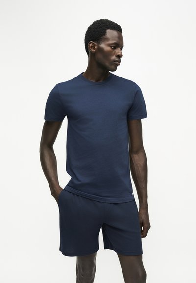 Homme aux cheveux courts et bouclés portant un t-shirt bleu marine uni et un short assorti, debout les mains dans les poches, regardant sur le côté.
