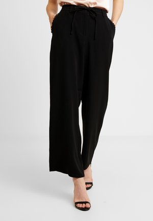 Femme portant un pantalon noir ample à cordon et des talons noirs ouverts à l'avant, les mains dans les poches, sur un fond clair uni.