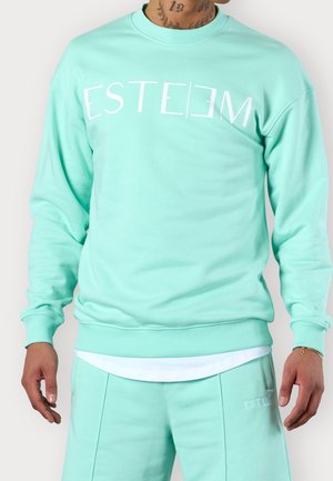Persona che indossa una felpa turchese e pantaloni abbinati con la parola "ESTEEM" stampata sul petto in lettere bianche.