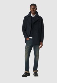 Granatowy wełniany płaszcz peacoat z podwójnymi guzikami, noszony na granatowym swetrze i koszuli z kołnierzykiem, w połączeniu z ciemnymi dżinsami i czarnymi butami.