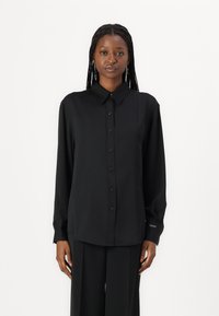 Camisa preta de botão com mangas longas, colarinho e uma textura suave. Apresenta um corte reto e detalhes discretos nos punhos.