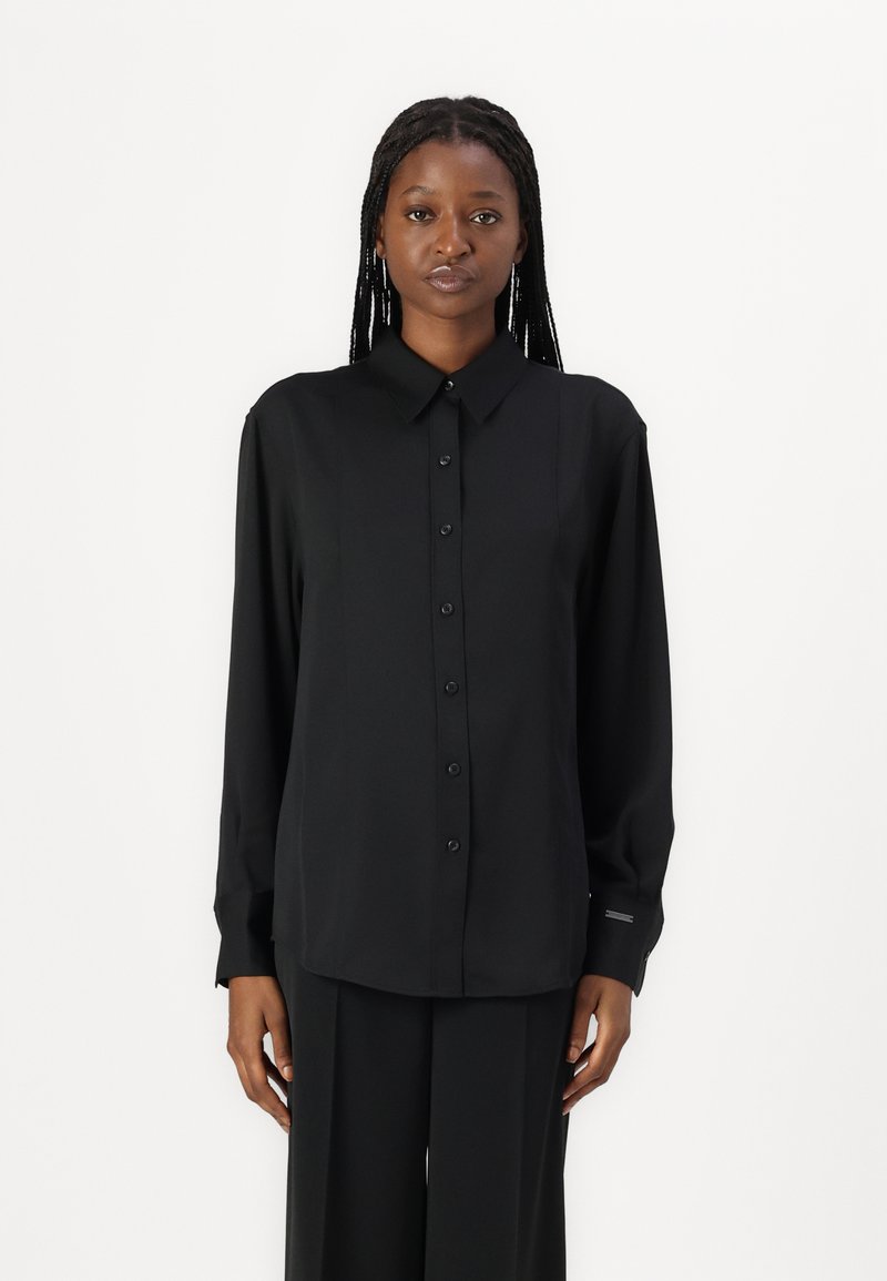 Camisa preta de botão com mangas longas, colarinho e uma textura suave. Apresenta um corte reto e detalhes discretos nos punhos.
