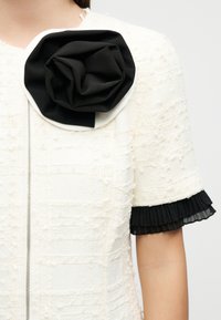 Λευκό φόρεμα με υφή, κοντά μανίκια, μαύρα πιετέ βολάν μανσέτες και ένα μεγάλο υφασμάτινο λουλούδι σε μαύρο και άσπρο καρφιτσωμένο κοντά στον λαιμό.