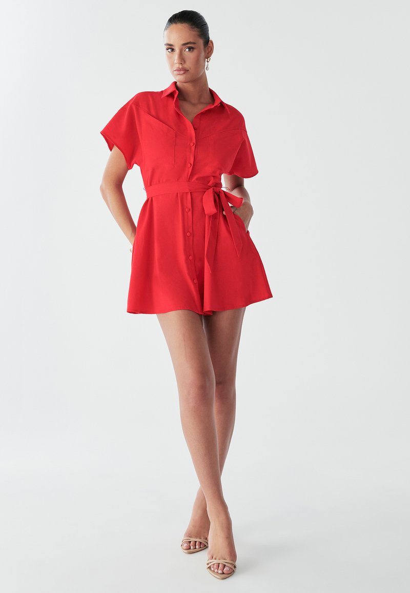 Model poseert in een kort rood jurkje met een strik in de taille, met een schone achtergrond en draagt beige sandalen met banden.