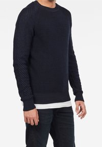 Marineblauer Pullover aus Strick mit strukturierten diagonalen Mustern an den Ärmeln, gerippten Bündchen und Saum, über einem weißen Hemd getragen. Weiches, leichtes Material.