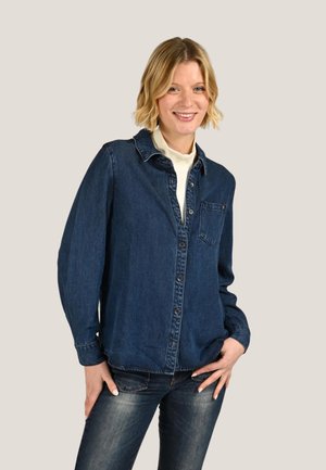 Femme souriante portant une chemise en jean foncée boutonnée sur un haut blanc, associée à un jean bleu délavé, debout, la main sur la hanche.