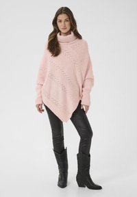 Ženska v roza pletenem ponču z vzorcem chevron, črnih usnjenih hlačah in črnih kavbojskih škornjih, ki stoji ob navadni ozadju.