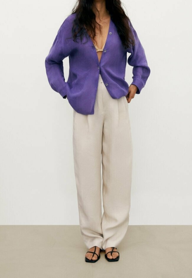 Cardigan violet oversize avec boutons à l'avant, porté sur une tenue beige clair. Un pantalon fluide beige et des sandales noires à brides complètent le look.