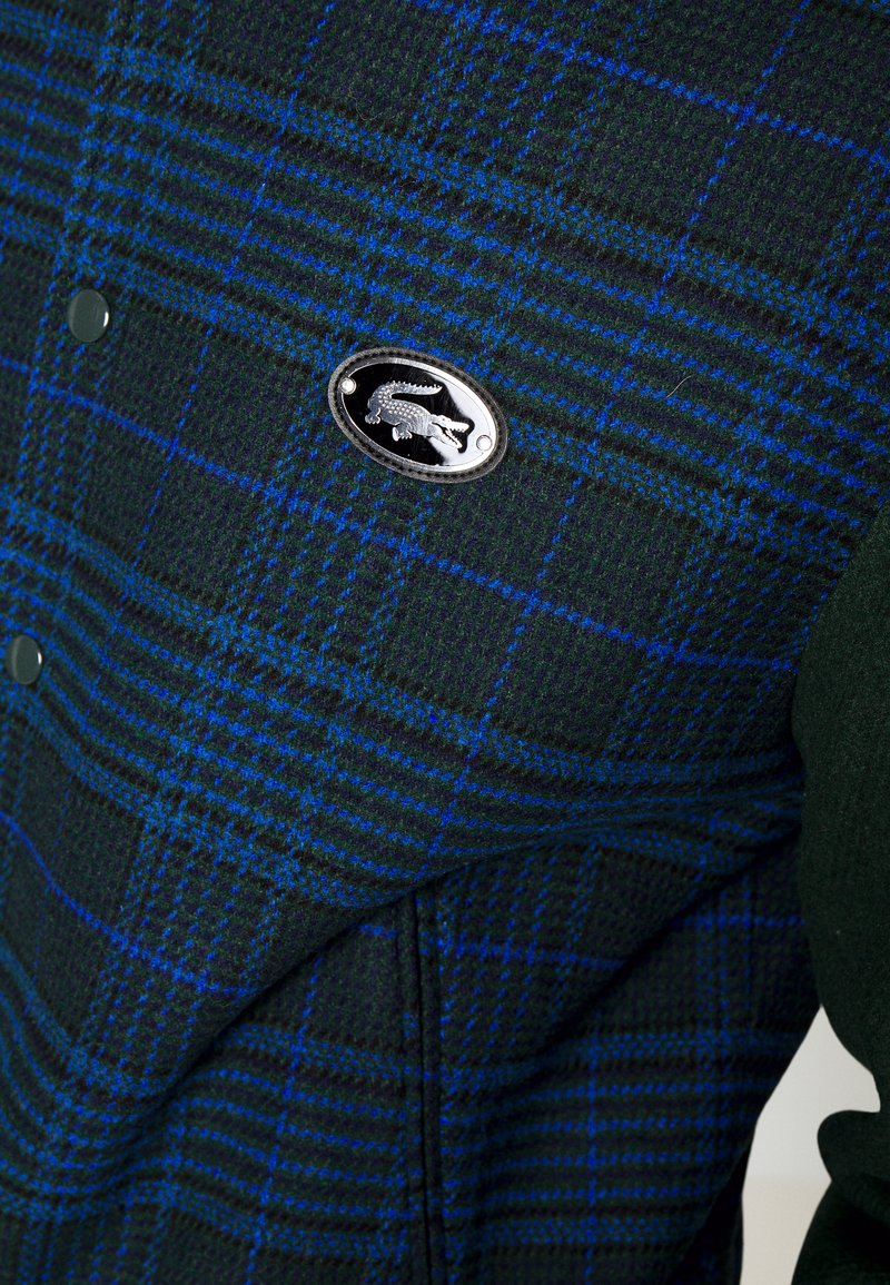 Veste à carreaux vert foncé et bleu avec des boutons noirs et un badge oval en métal argenté en forme de crocodile sur la poitrine.