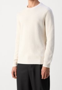 Cremefarbener Pullover aus Rippenstrick mit Rundhalsausschnitt, langen Ärmeln und einem enganliegenden, gerippten Saum, getragen über einer dunklen Hose.