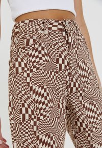 Bruine en witte geruite broek met een draaiend geometrisch patroon, voorzien van metalen knoopsluitingen en zijzakken. Gelaagde stof.