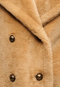 Μπεζ μαλακό παλτό από ψεύτικη γούνα με φαρδιά γιακά και τρία ορατά σκοτεινά μεταλλικά κουμπιά σε στενή όψη.