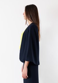 Giacca blu navy dalla vestibilità ampia con maniche larghe, indossata sopra un top giallo brillante. Tessuto liscio, design minimalista, senza chiusure visibili.