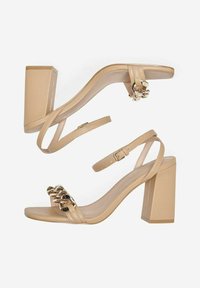 Las sandalias de tacón alto beige cuentan con un tacón de bloque, una correa de tobillo ajustable y un acento de cadena de metal en la correa del dedo del pie, con una textura suave.