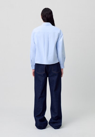 Chemise bleu clair à manches longues avec des poignets à boutons et un pli au dos, associée à un jean à jambes larges bleu foncé, vue de dos.
