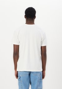 Man som står med ryggen mot kameran, iklädd en enkel vit t-shirt med kort ärm och ljusblå denim cargo-byxor, mot en vit bakgrund.