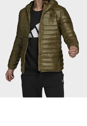 Homme portant une doudoune vert olive par-dessus un t-shirt Adidas noir et un pantalon noir, posant avec les bras légèrement étendus.