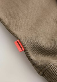 Kaki tröja med texturerat stickat tyg och ribbad mudd. Har en ljus röd etikett med "SUPERDRY"-logotyp på sidan.