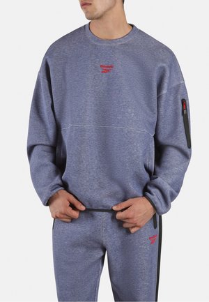 Mann trägt blaues Reebok-Sweatshirt und passende Jogginghose, Hände justieren den Bund, rote Reebok-Logos auf Brust und Hose.