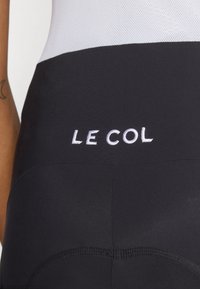 Black cycling shorts made of smooth fabric; features white embroidered "LE COL" logo, flat seams, and a contoured design for comfort.